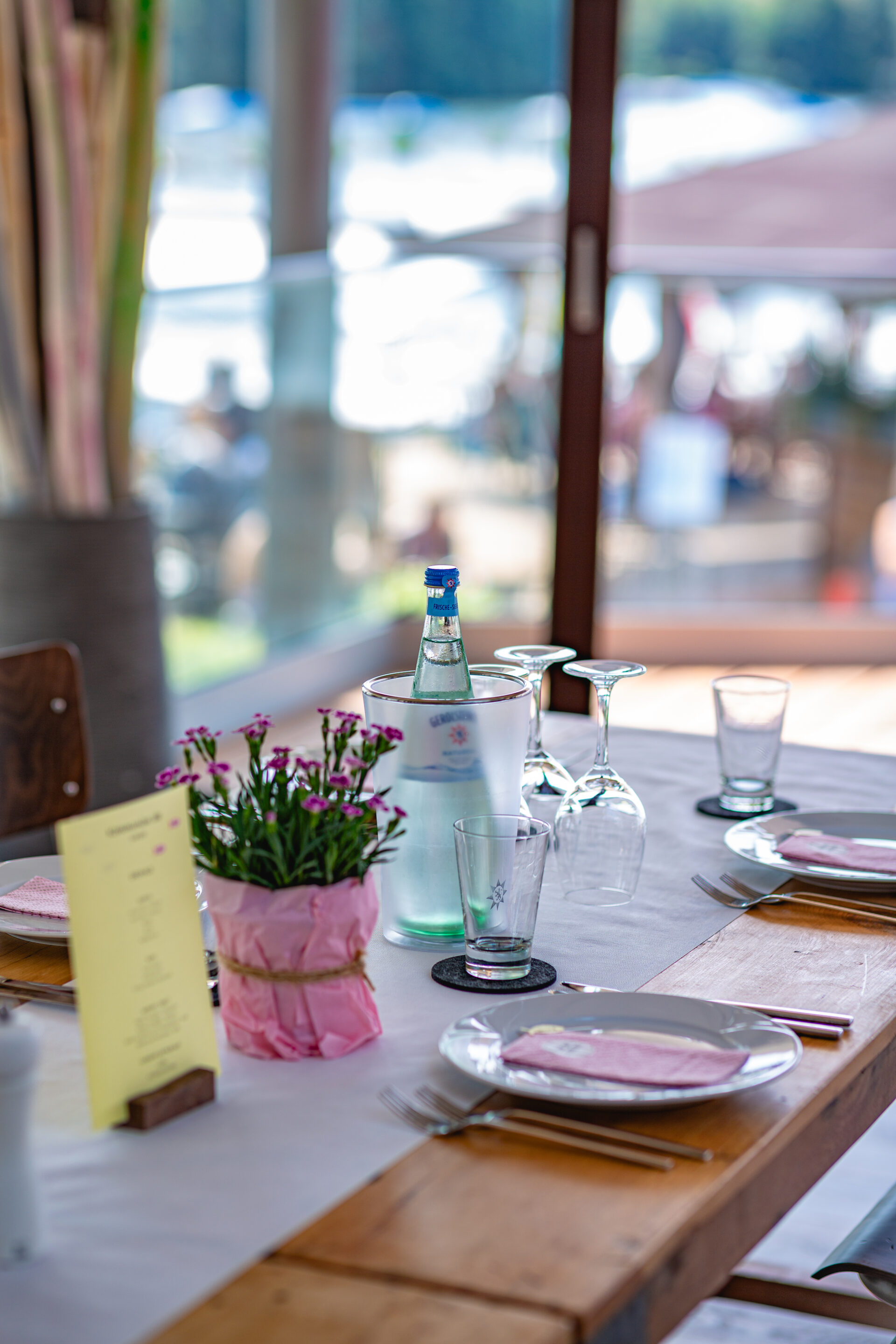 Eingedeckter Tisch im Eventraum mit weißen porzellan Tellern, rosa Serviette, Blumen, Wasser und Gläsern.
